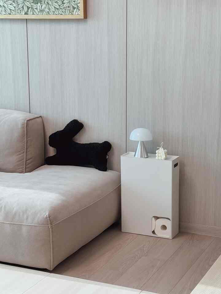 明るいグレーの壁と木目調の床のリビング空間、ベージュのソファに黒いうさぎクッション、白のスリムなトイレットペーパー収納が配置されたモダンで洗練されたインテリア