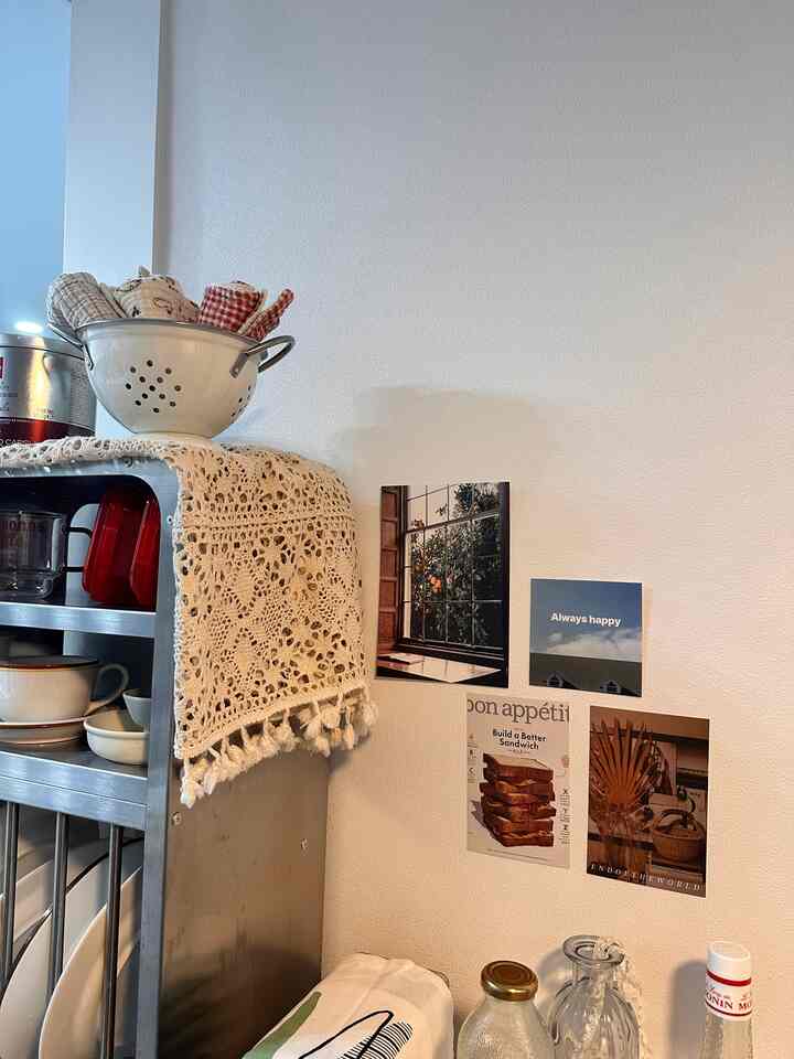 白い壁にヴィンテージポスターとクロシェテーブルクロスが飾られたキッチンの居心地の良いおうちカフェ空間