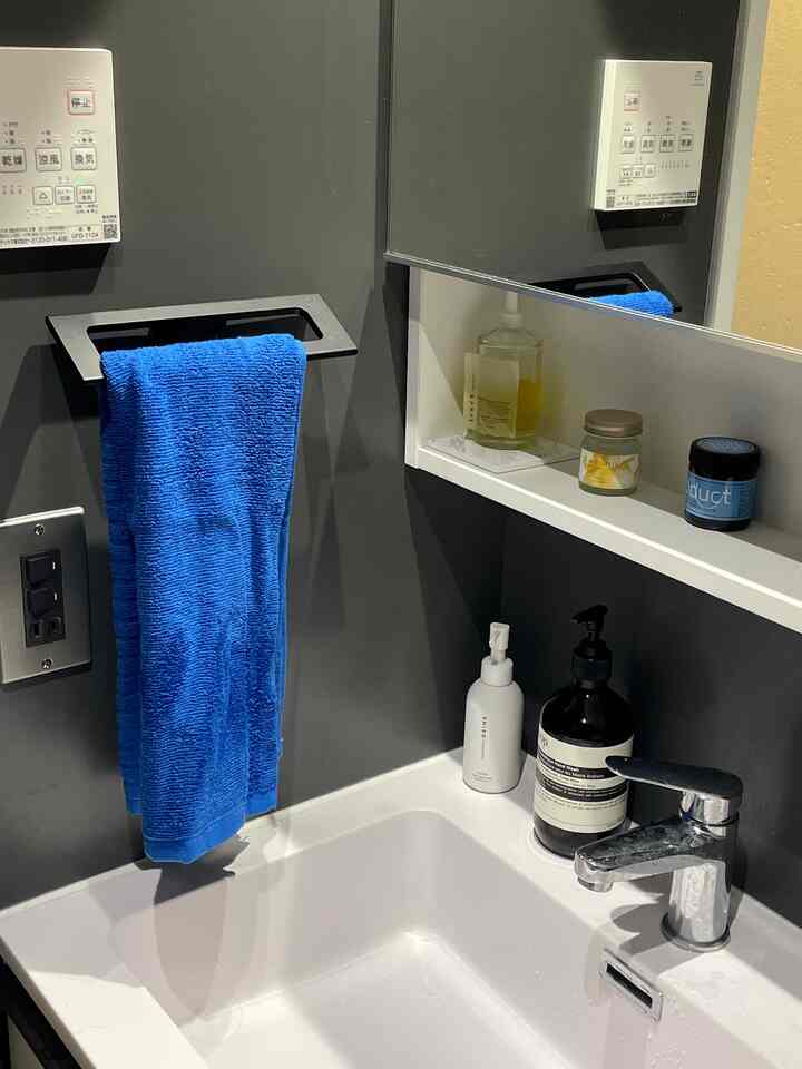 グレーの壁と白い洗面台があるモダンなバスルーム、青いタオルが掛かりイソップのハンドウォッシュが整然と配置された清潔感のある空間