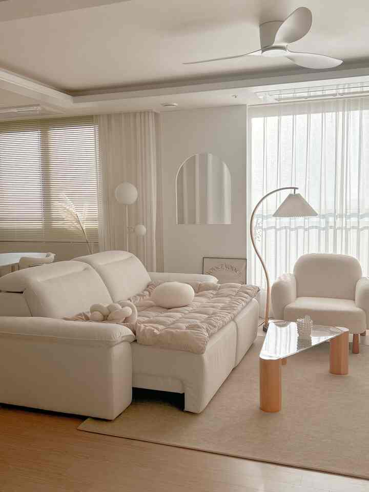 白とベージュのトーンのリビングルーム、クリーム色のソファとクッション、三角形のコーヒーテーブル、明るいカーテンとフロアランプが配置された雰囲気のある空間