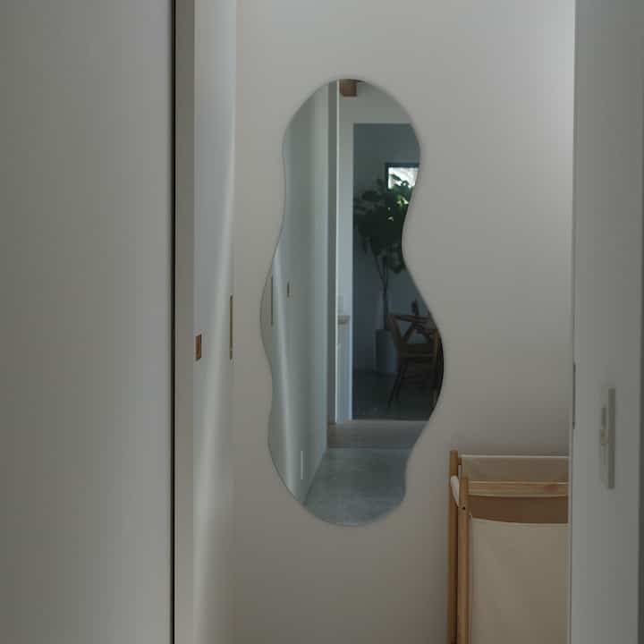 柔らかな曲線の鏡が中央に掛かった白い壁とナチュラル素材のランドリーバスケットがあるシンプルな玄関の雰囲気のあるインテリア