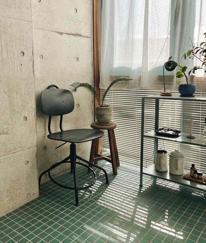 グリーンタイルの床とコンクリートの壁が調和した狭い空間、ブラックチェアと木製スツール、棚ユニットが温かな自然光の中に配置されたインダストリアルな雰囲気のインテリア