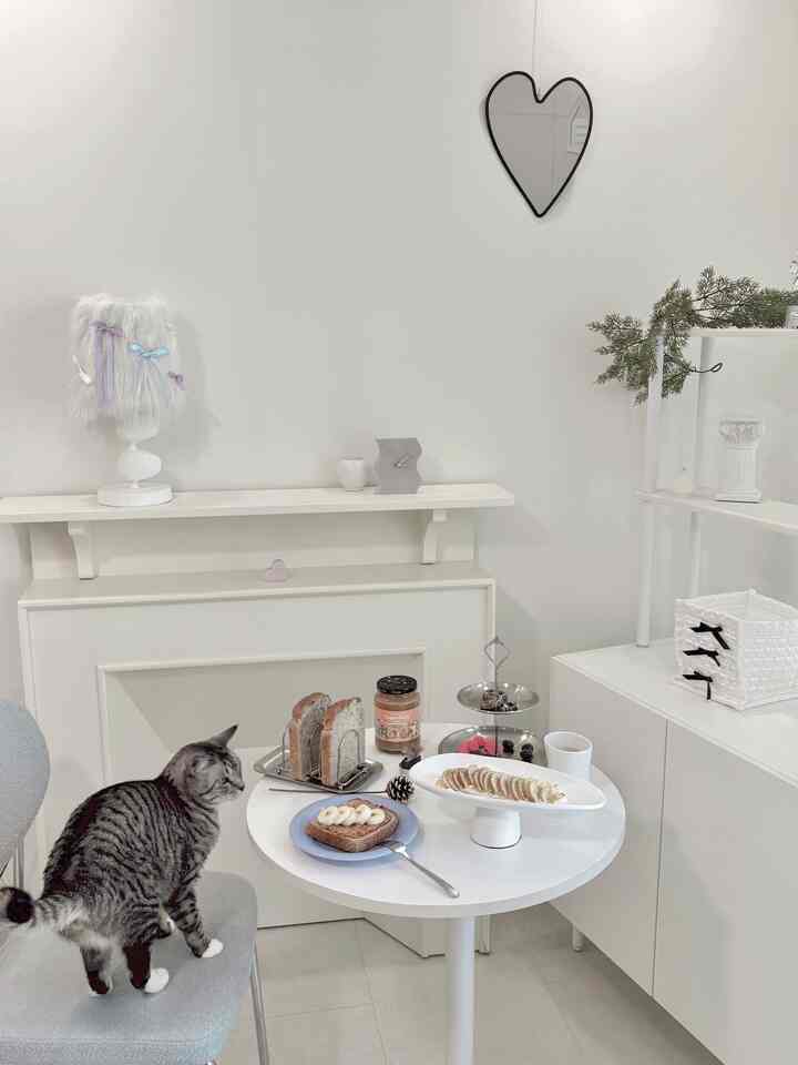 ホワイトトーンのかわいらしいおうちカフェ、青い椅子に猫が座り、小さな丸テーブルにトーストとデザートが置かれた雰囲気のあるインテリア