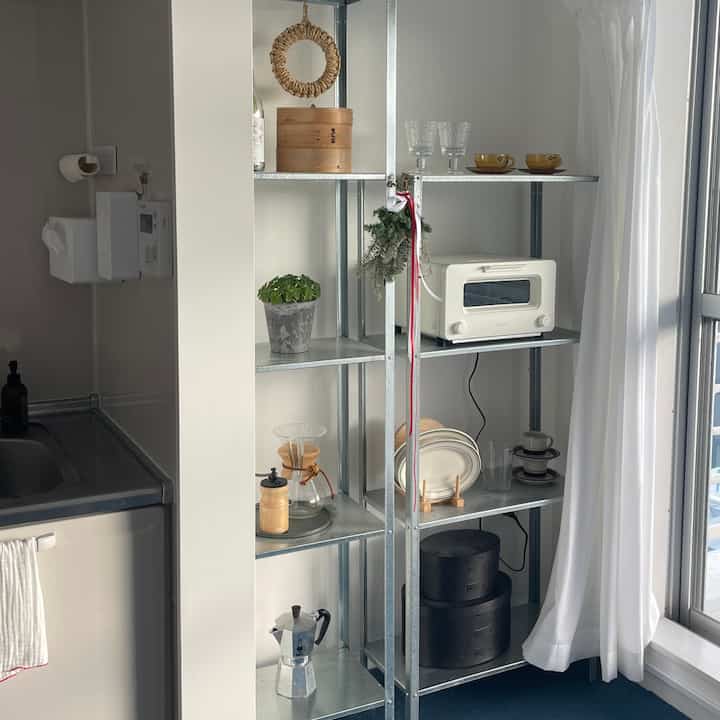 Light-filled small kitchen with white walls and curtains, metal shelving organized with coffee equipment and kitchenware, creating a cozy vibe