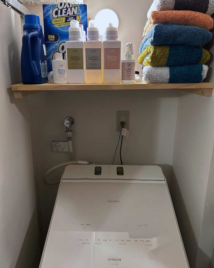 ナチュラルカラーの壁と白い洗濯機、その上にタオルと洗濯用品が整頓された木製棚が配置されたコンパクトなランドリールーム