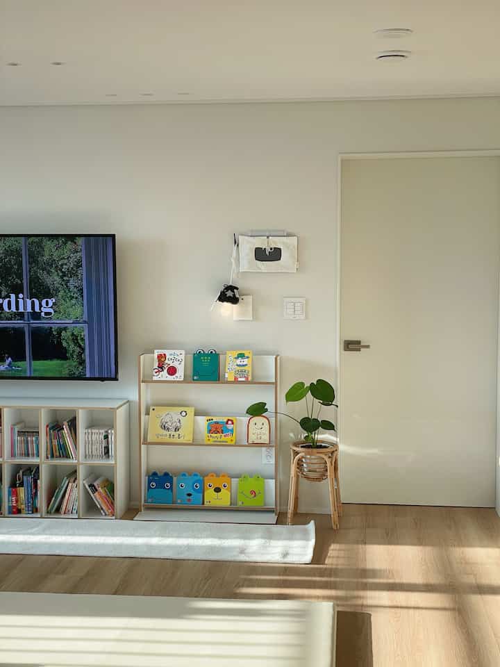 Bright natural-toned living room featuring children's bookshelves and a plant stand, creating a warm and tidy atmosphere