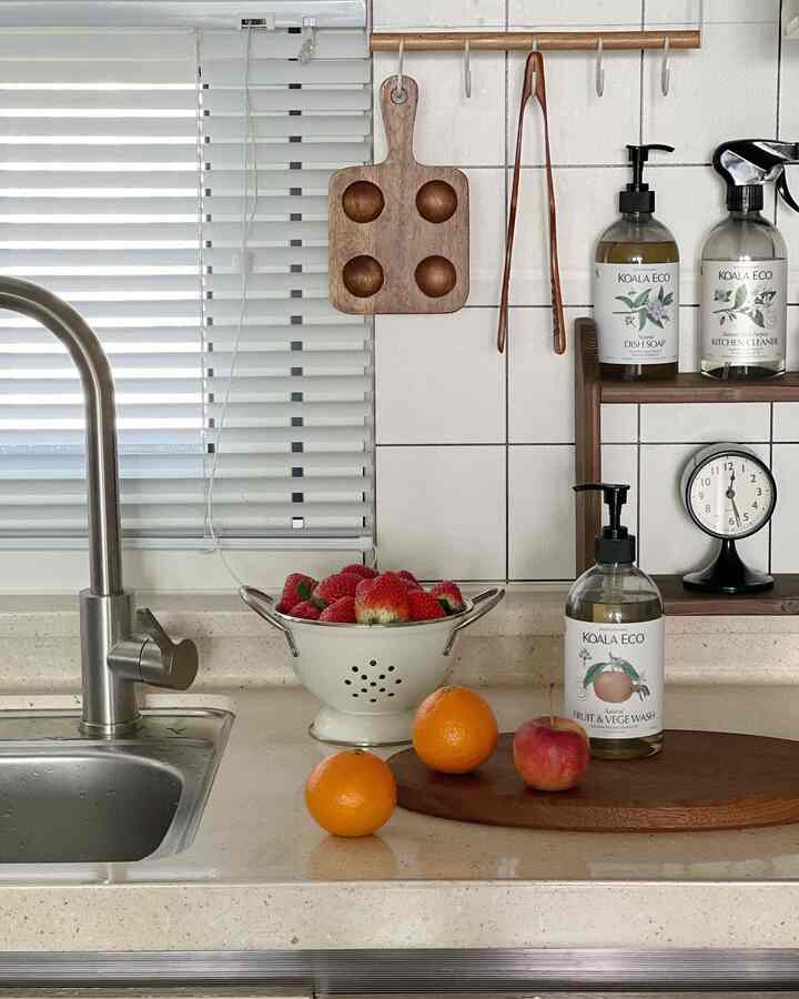 白と木目調のキッチン、ブラインドとシンク、新鮮な果物と調理用品が配置された清潔感のある空間