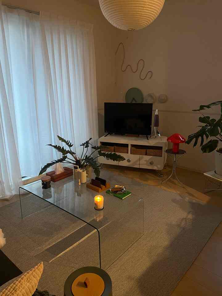 白いカーテンとナチュラルトーンの床が調和するリビング、透明なガラスのコーヒーテーブルと植物が配置された雰囲気のある空間