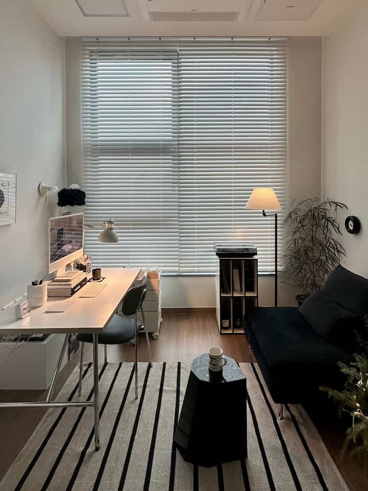 A white and black toned studio home office featuring velvet sofa, wood blinds, and a striped rug creating a cozy atmosphere.