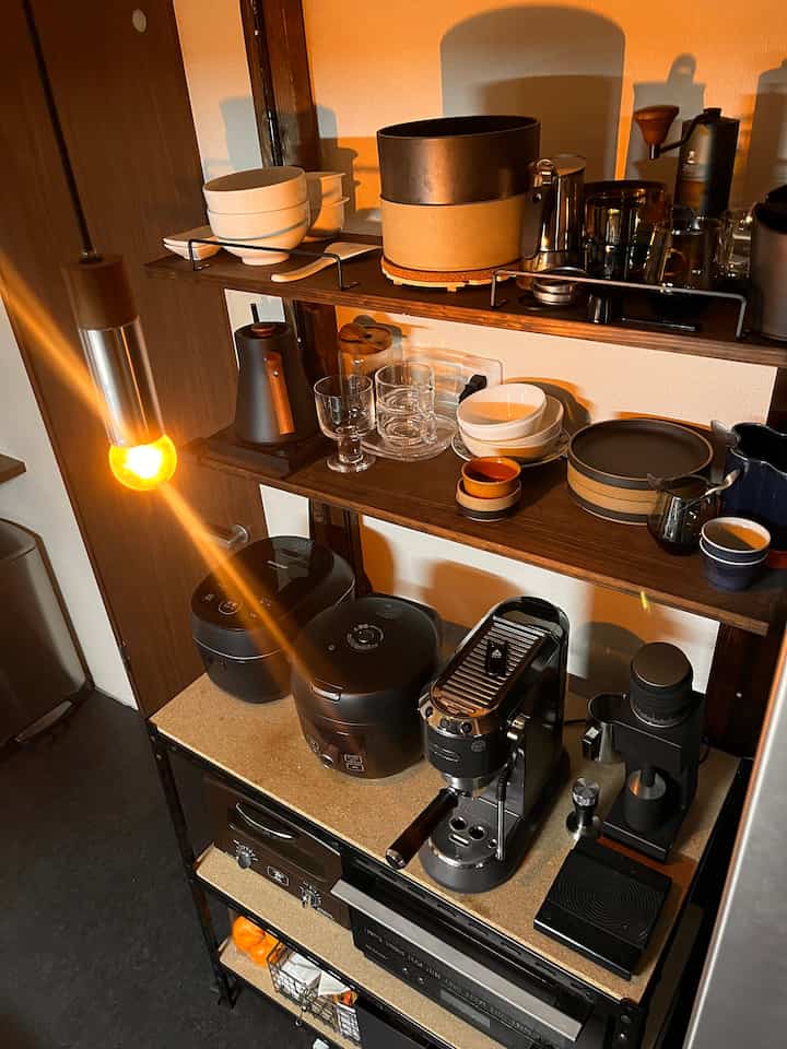 暖かい照明があるナチュラルな色調のキッチン収納棚にコーヒーマシンやグラインダーなどホームカフェ用品が配された印象的な空間