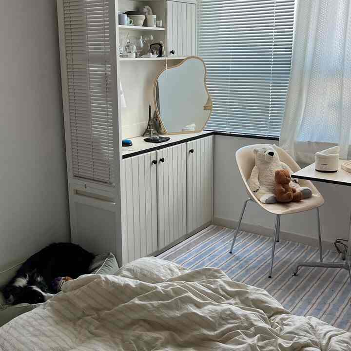 白とグレーの色調の寝室、ストライプのラグ、ブラインド、不規則な形の鏡が配置されており、落ち着いた自然な雰囲気のあるインテリア