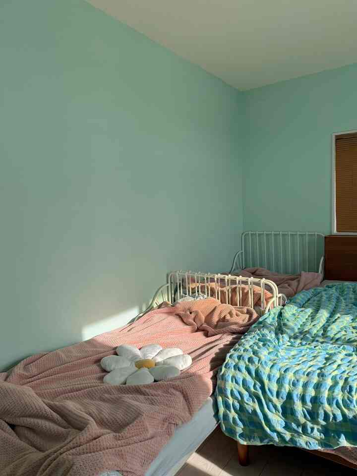 ミントグリーンの壁と自然光が映える小さな子供部屋、ピンクとブルーの掛け布団が掛けられた二つのベッドを配した温かみのあるインテリア