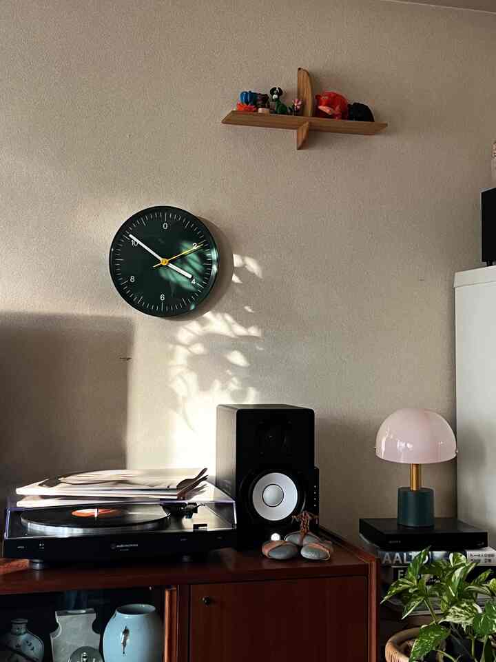 Natural-toned living space corner with green clock, wooden shelf, record player, and speaker showcasing classic interior style