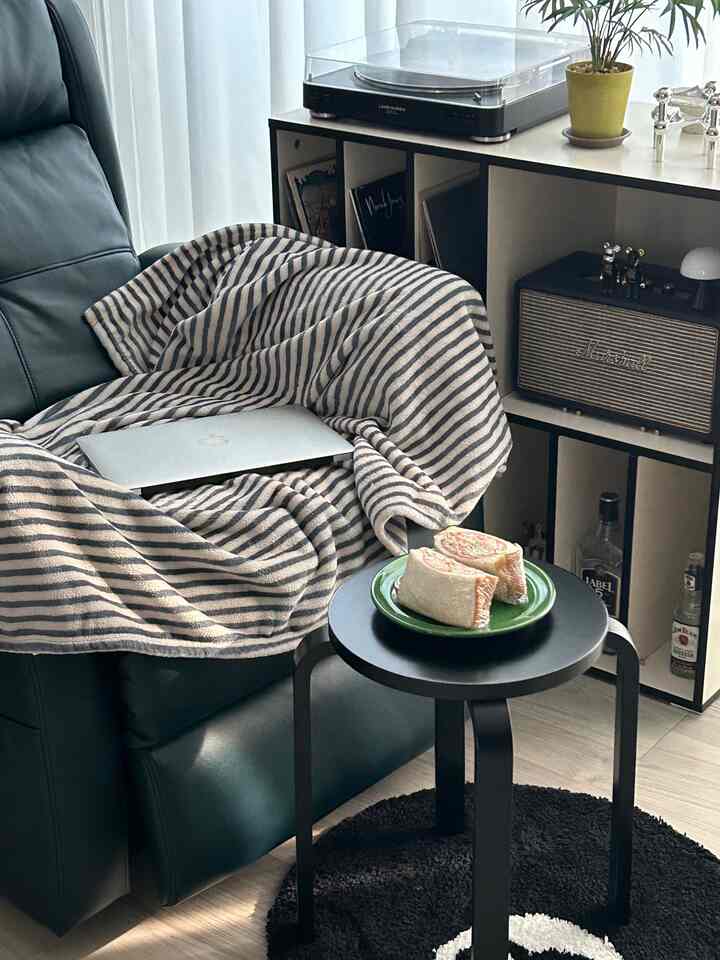 ブラックとグレーのトーンのリビング、リクライナーとストライプのブランケット、丸いスツールの上にクラミとキュウリのサンドイッチが置かれた温かみのある空間