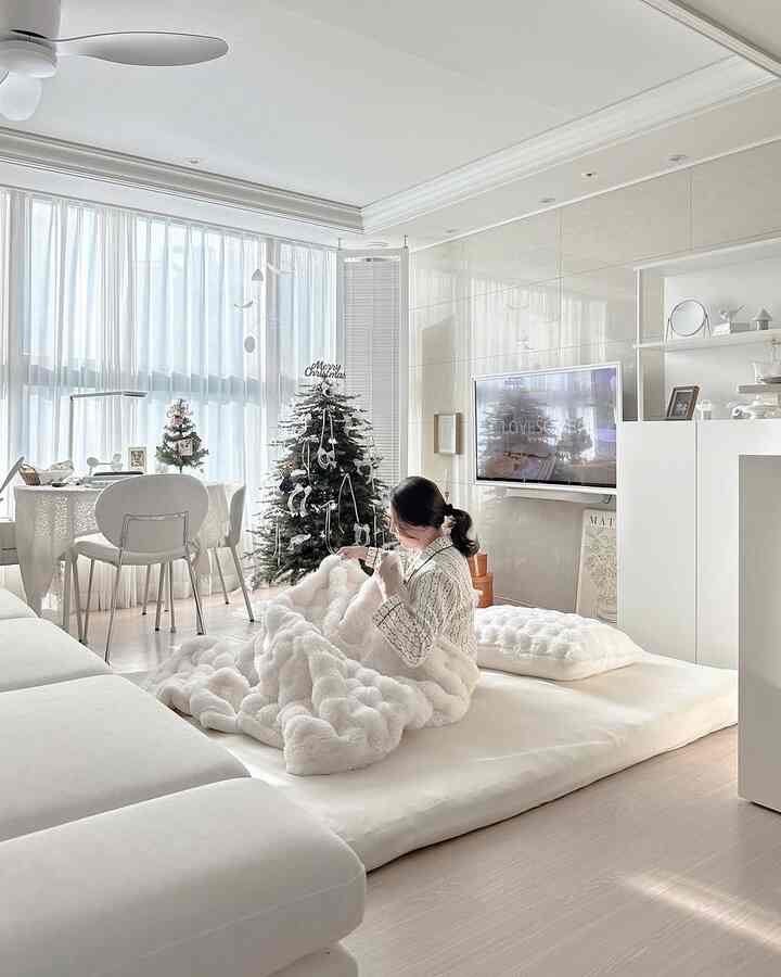 明るいホワイトトーンのリビングルーム、ソファとカーテンの前に温かみのあるミンクファーブランケットに包まれた人が座るミニマルで冬らしい空間