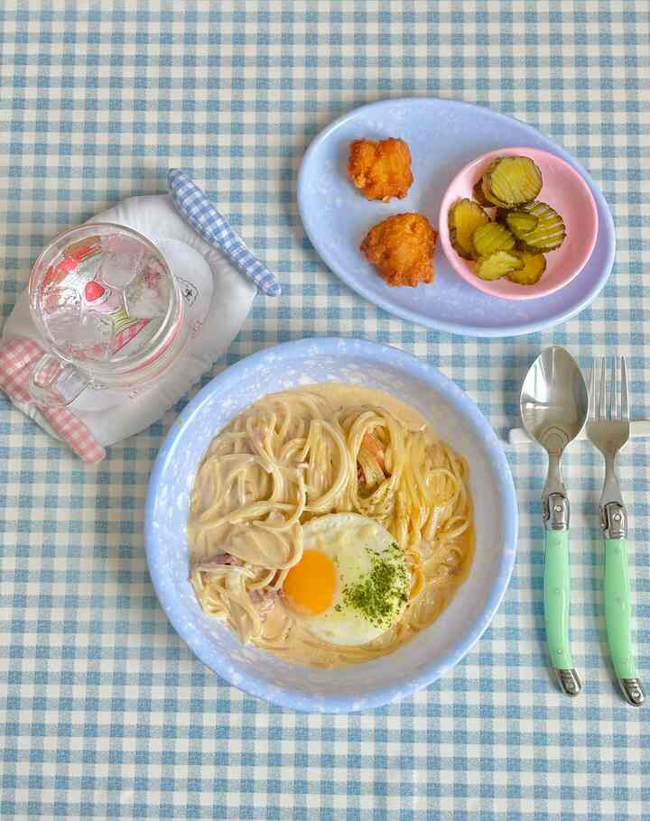 Pastel-toned blue checkered tablecloth with creamy pasta, pink and blue tableware, and mint cutlery setting a cozy dining atmosphere