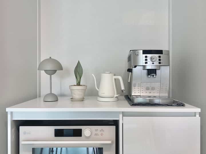 White and silver toned kitchen area featuring a coffee machine, electric kettle, small plant, and table lamp creating a clean, modern home cafe space