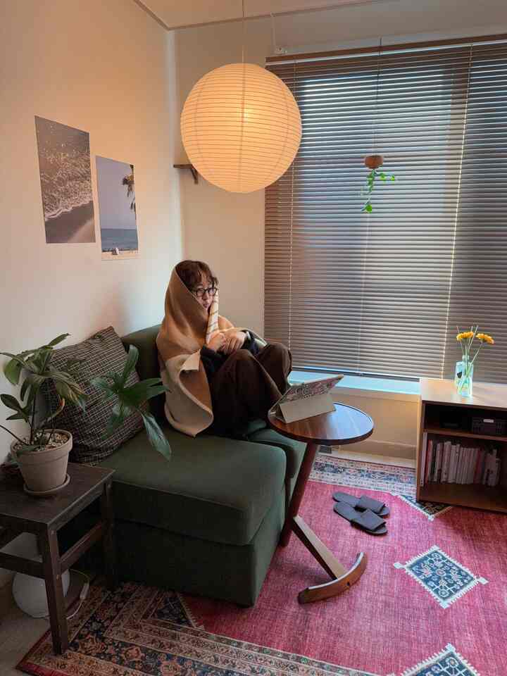 柔らかなブラウンとグリーンの色調が特徴のリビング、ソファに腰掛けてブランケットを羽織る人がいる雰囲気のあるホームオフィス空間