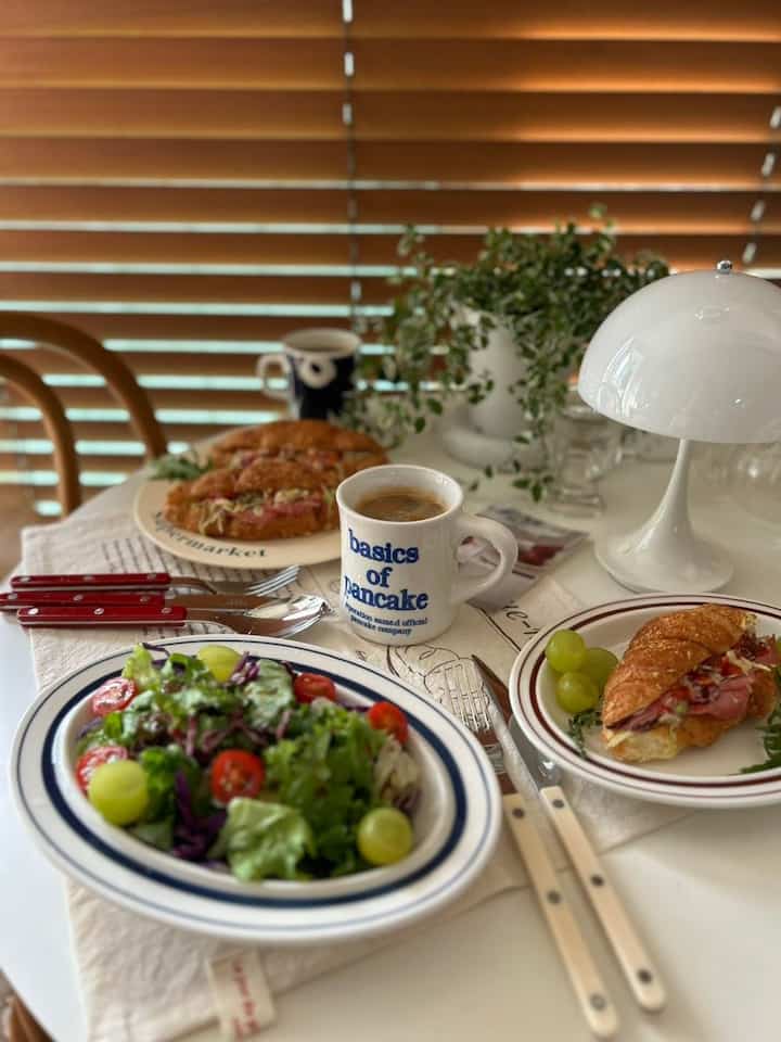 White dining table with brown wooden blinds and chair, featuring salad, croissant, and coffee in a cozy home cafe brunch setting