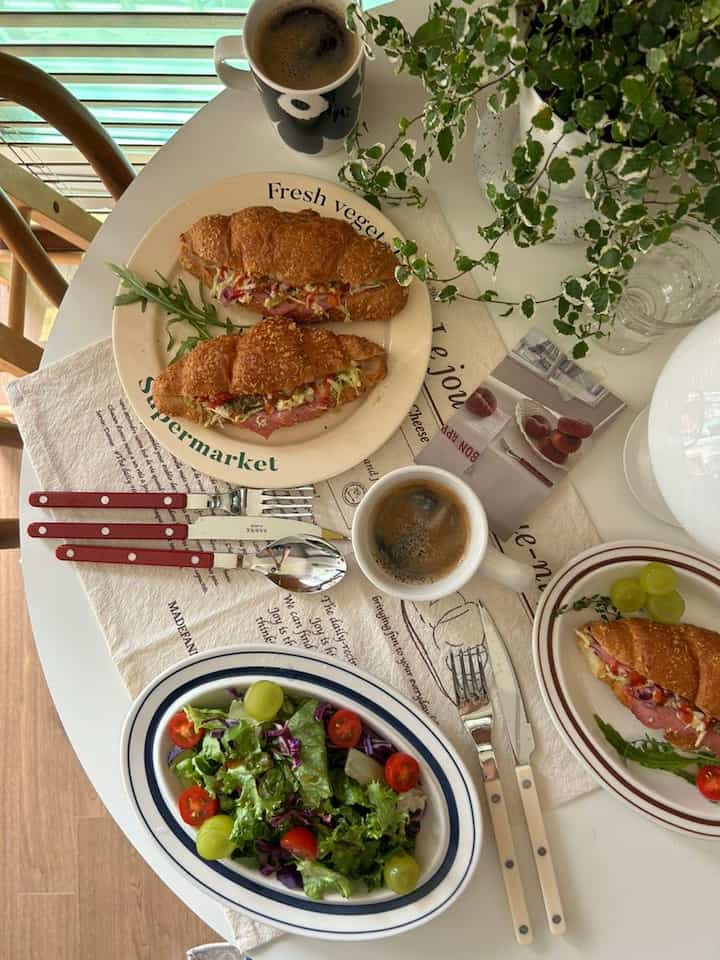 White round dining table with croissant sandwiches, salad, coffee cups, and plant creating a cozy cafe style home cafe atmosphere