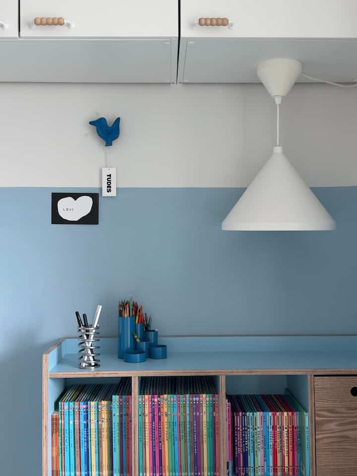 A kids' room in white and blue tones featuring a bookshelf and pendant light creating a neat and cozy atmosphere