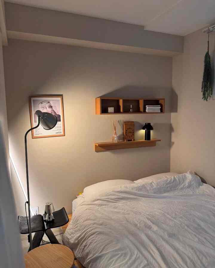 ベージュの壁と白い寝具が調和するシンプルモダンな寝室、天然木の棚と黒いフロアランプが配置された雰囲気ある空間