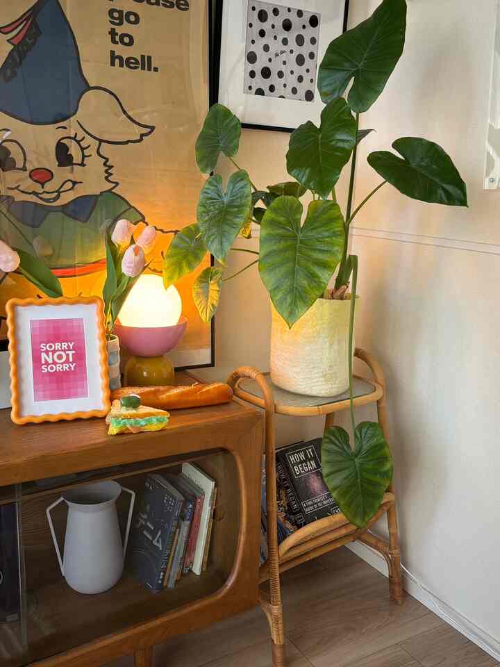 暖かみのある木目調と間接照明が調和したリビングの一角、ラタンのプラントスタンド、額縁、ポスター、大きな観葉植物が配置された印象的な空間