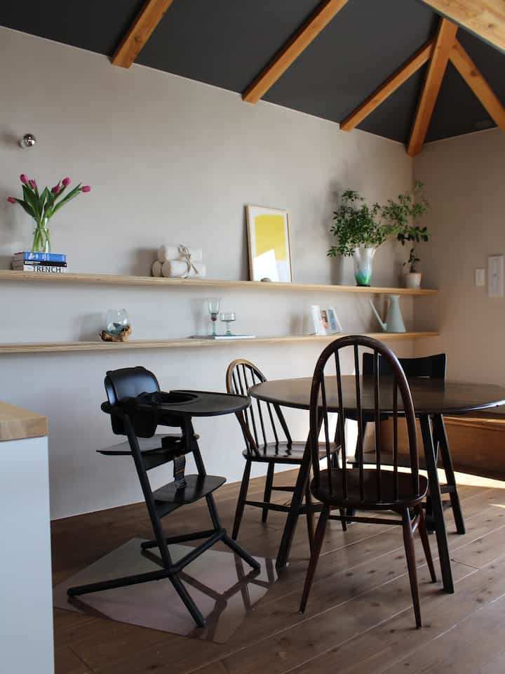 A beige-walled dining room with wood tone flooring featuring a round dining table, vintage chairs, and a baby high chair creating a natural space