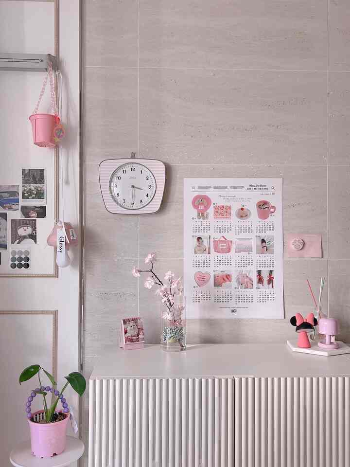 Simple interior space with soft white and pink tones featuring a clock, poster, plant, and pink decor in front of a living room wall