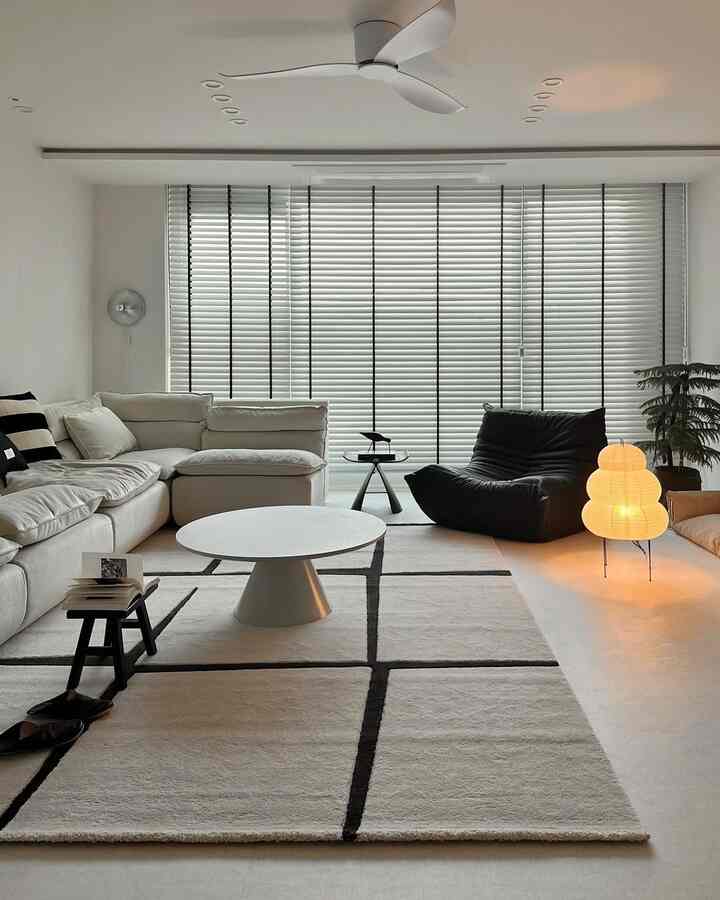 Monotone living room featuring a white sectional sofa, a round coffee table, large blinds, and a warm glowing floor lamp in a modern setting