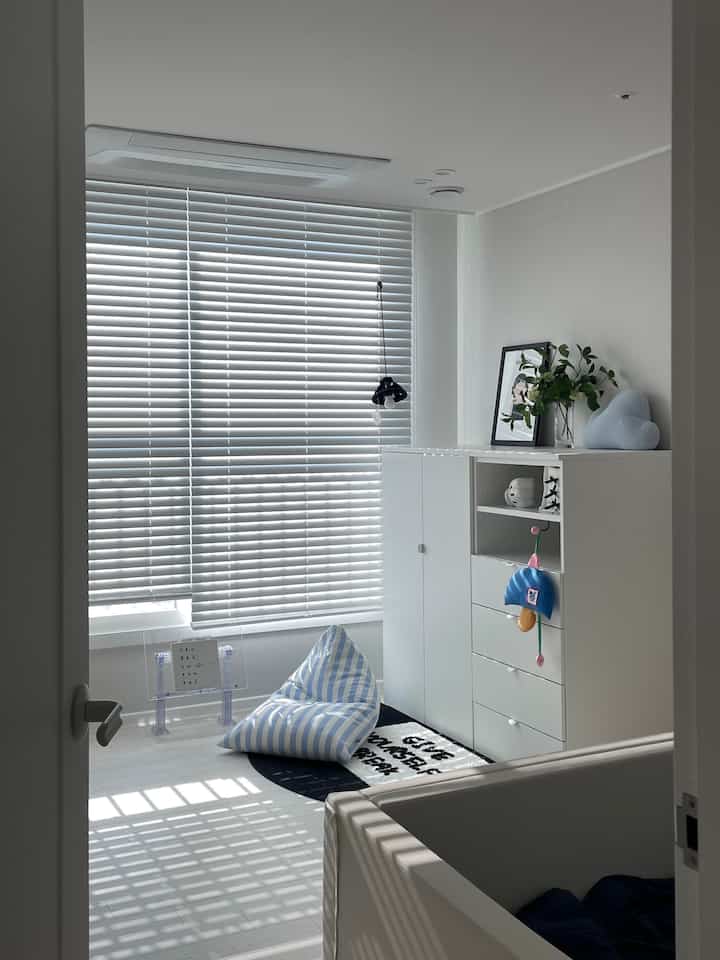 白と青の明るい子供部屋、ベビーベッドとビーンバッグ、ブラインドが配置された清潔感のある印象的な空間
