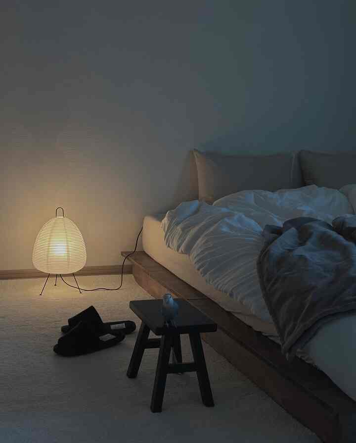 A wood tone-dominant low platform bedroom featuring white bedding, black stool, and a soft mood lamp creating a cozy atmosphere
