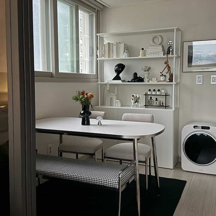 白と黒のトーンが特徴的なワンルームのリビング兼ダイニングスペース、清潔感あるモダンなダイニングテーブルとベンチ、棚が配置されている印象的な空間