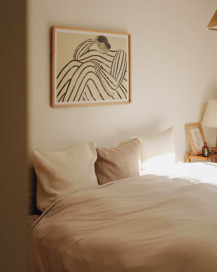 アイボリー調の6畳寝室、クイーンベッドと枕、壁にかかったミニマルなウォールアートが調和するナチュラルな雰囲気のインテリア