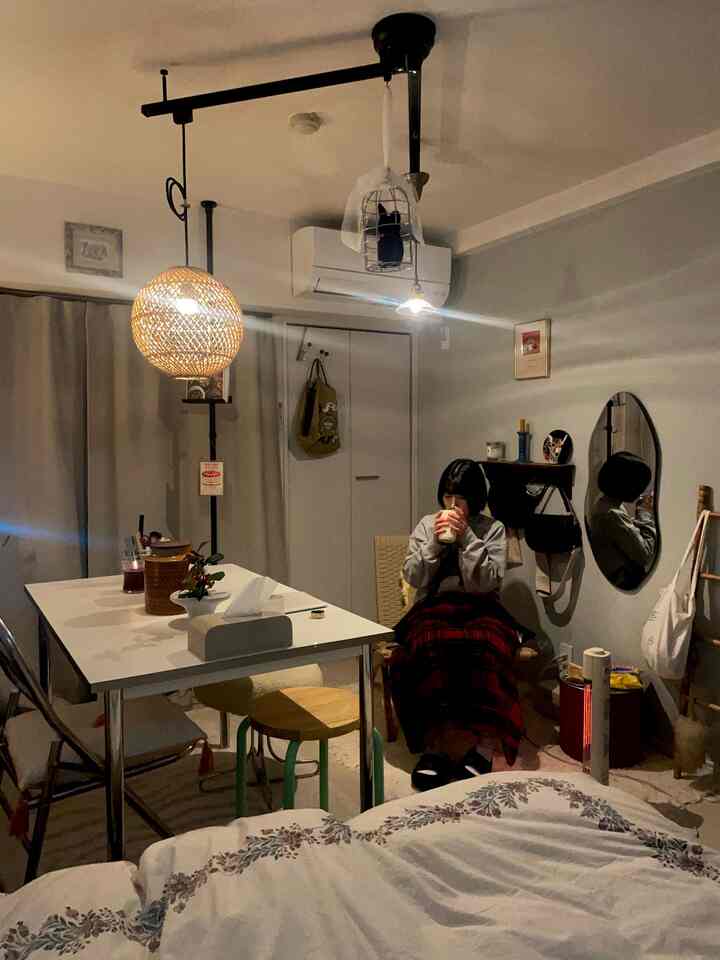 ダークグレーのトーンが特徴の1K一人暮らしリビング、白いテーブルと木製スツール、暖かいペンダントライトの下でチェック柄の毛布に包まれた人が座っている雰囲気のあるインテリア