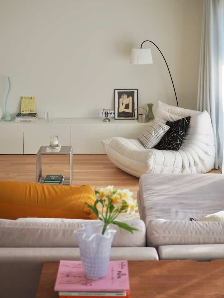 明るいベージュとブラウンのトーンのリビング、中央にナチュラルホワイトのソファとフロアランプ、手前に透明な花瓶とオットマンが配置された居心地の良い空間