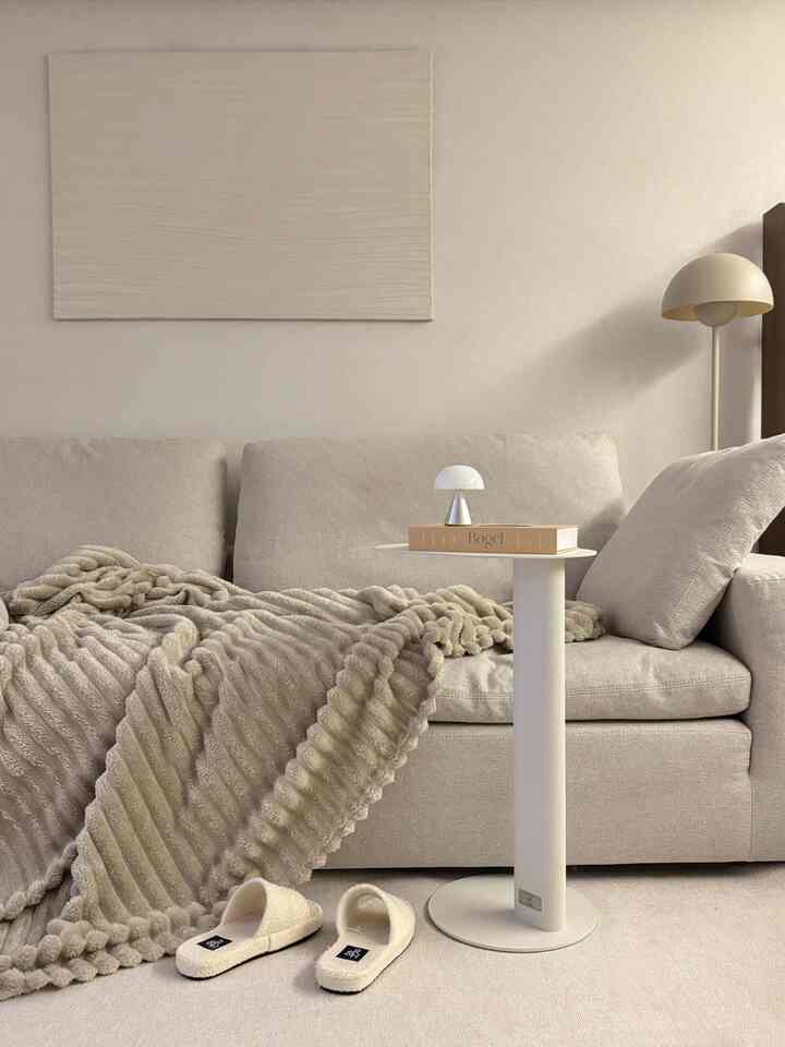 Beige and natural tone living room featuring a beige sofa, round white side table, cozy blanket, and slippers centrally arranged
