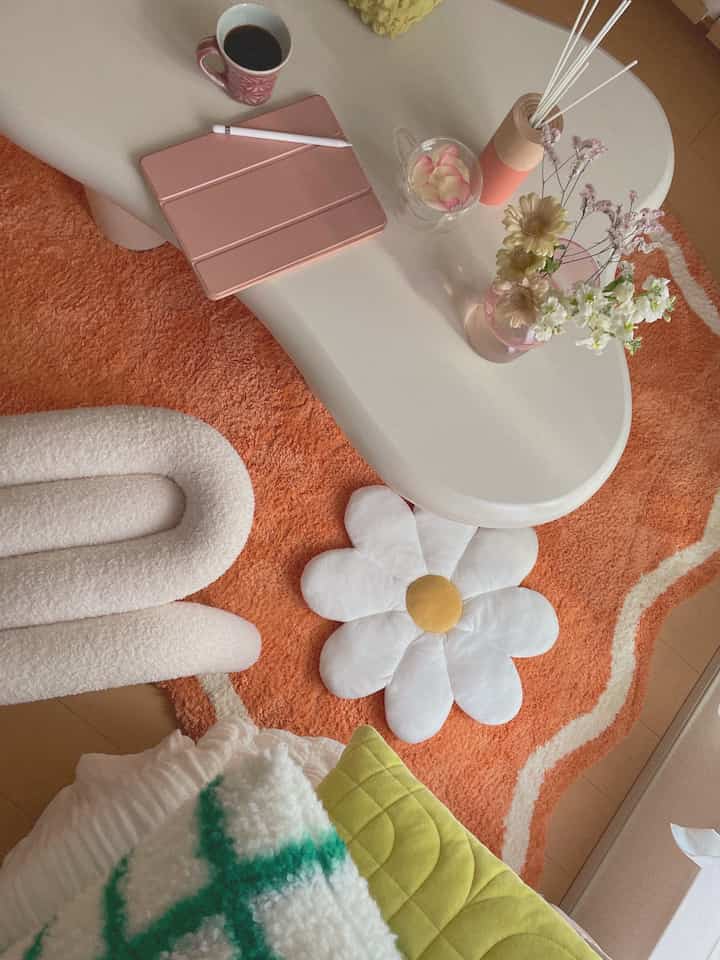 暖かいオレンジのラグと白いテーブルがあるカフェ風の空間、花瓶とクッションが配置されていて居心地の良い雰囲気を演出している