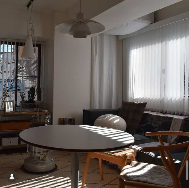 明るいホワイトと木目調が調和するリビング、バーチカルブラインド越しの自然光とラウンドテーブルを囲む暖かみのある家具のある印象的な空間