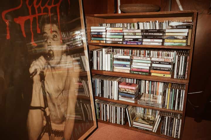 ブラウンの木製棚に色とりどりのCDとカセットテープ、大型ポスターが配置された音楽スペース
