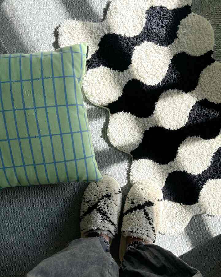 Monotone and light gray bedroom space featuring a monochrome patterned rug, green check cushion, and soft suede-like slippers creating a cozy atmosphere