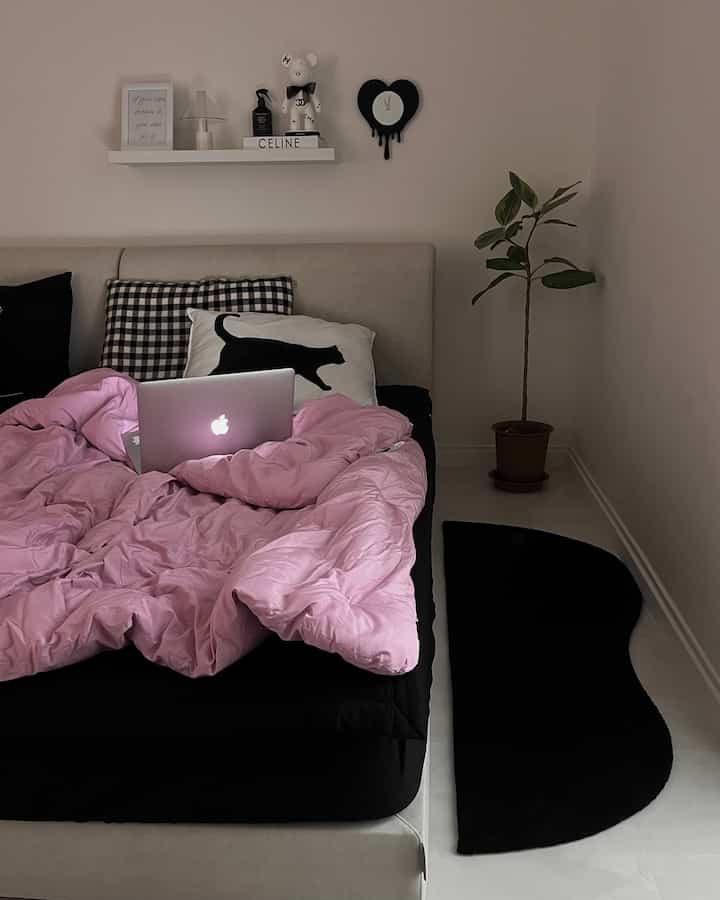 ピンクの寝具と黒のベッドベースが特徴のモダンな寝室、整然とした棚と観葉植物が置かれた居心地の良い空間