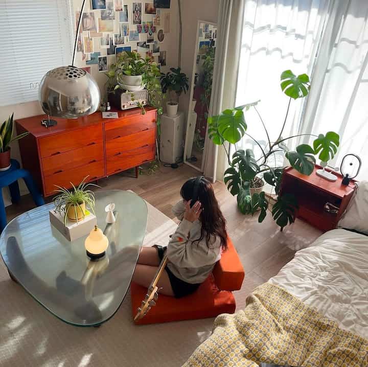温かみのある木目調と緑の植物が配された8畳の寝室、オレンジの座椅子と透明な三角形のテーブルが配置された雰囲気のある空間