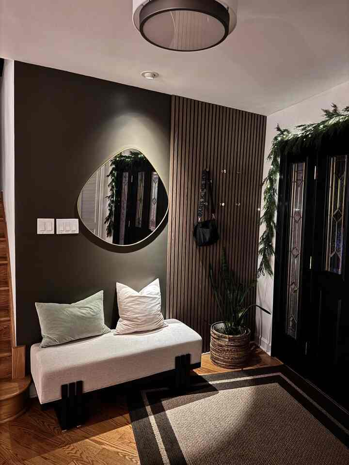 Modern entrance with charcoal wall and wood tone slat panel, featuring a cozy bench and uniquely shaped wall mirror creating a welcoming atmosphere
