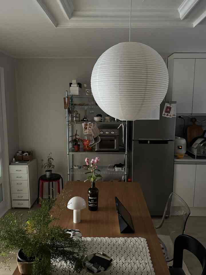 ナチュラルカラーのキッチン、中央に大きなペンダントライトと木製のダイニングテーブル、透明な椅子と緑の植物が配置された雰囲気のあるインテリア