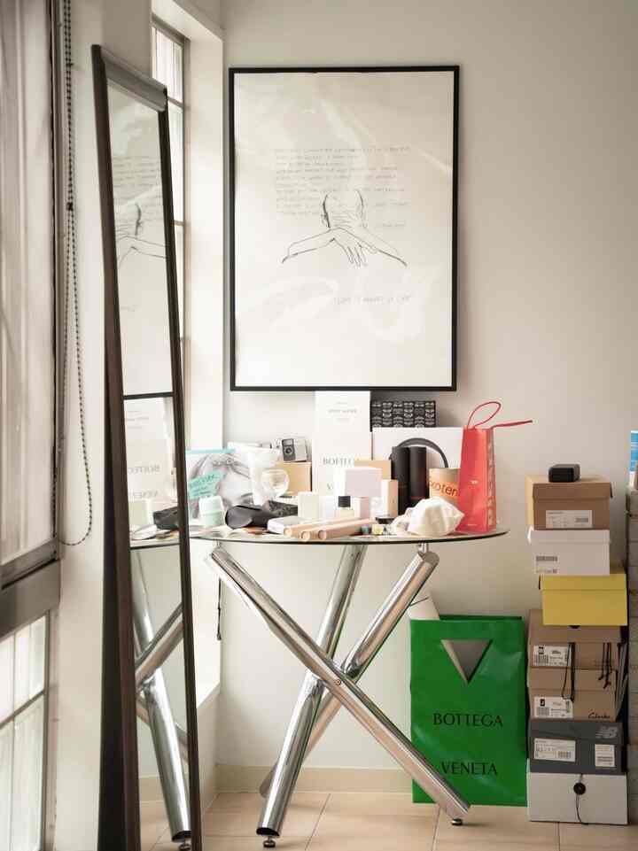 A small room with white walls and brown floor featuring a glass table with aluminum legs, a standing mirror, and stacked boxes with various items in a modern interior