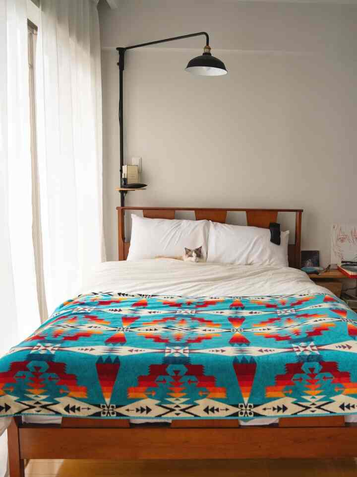 Natural and white tone bedroom featuring wooden bed with white bedding, vibrant patterned blanket, and a cat resting on the bed, creating a cozy atmosphere