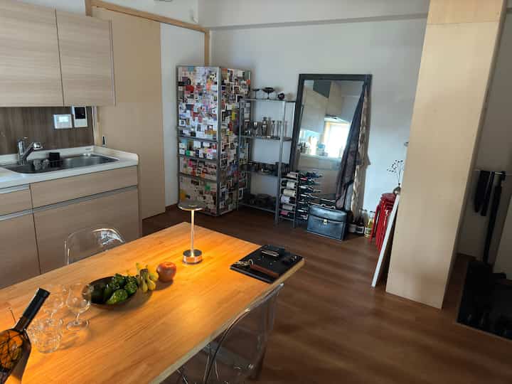 ナチュラルな木目調の1LDKの玄関兼ダイニングルーム、木製の食卓とスツール、冷蔵庫、ワインラックが配置された落ち着いた空間