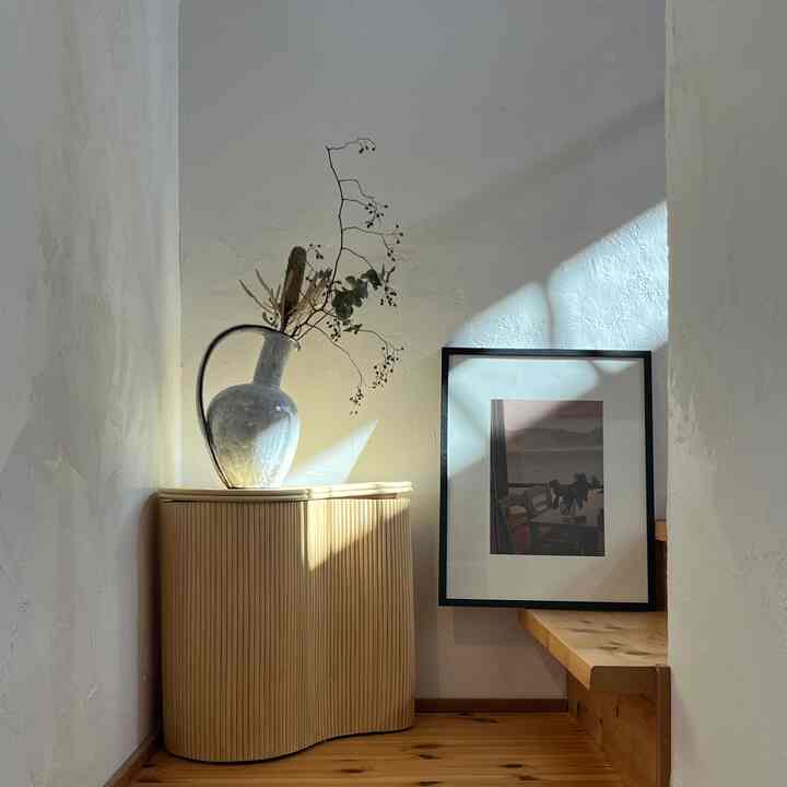 Natural style stair corner with white walls, wood tone floor, a corner storage side table featuring a ceramic vase, and framed poster creating a cozy atmosphere