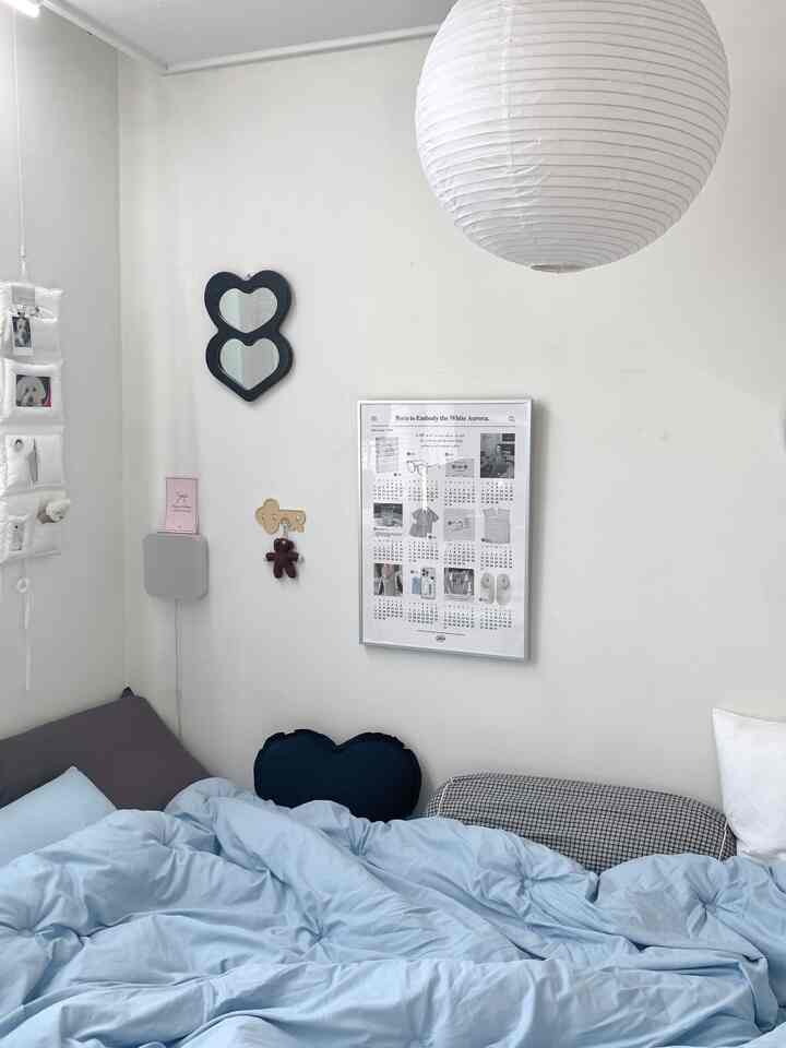 白い壁と青い寝具が配置された小さな寝室、ハート型クッションとヴィンテージ風の壁掛けミラーが印象的な空間